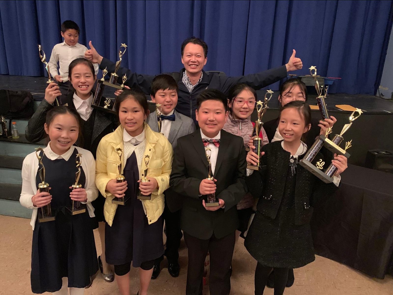 A squad of elementary schoolers holding trophies in front of a blue curtain